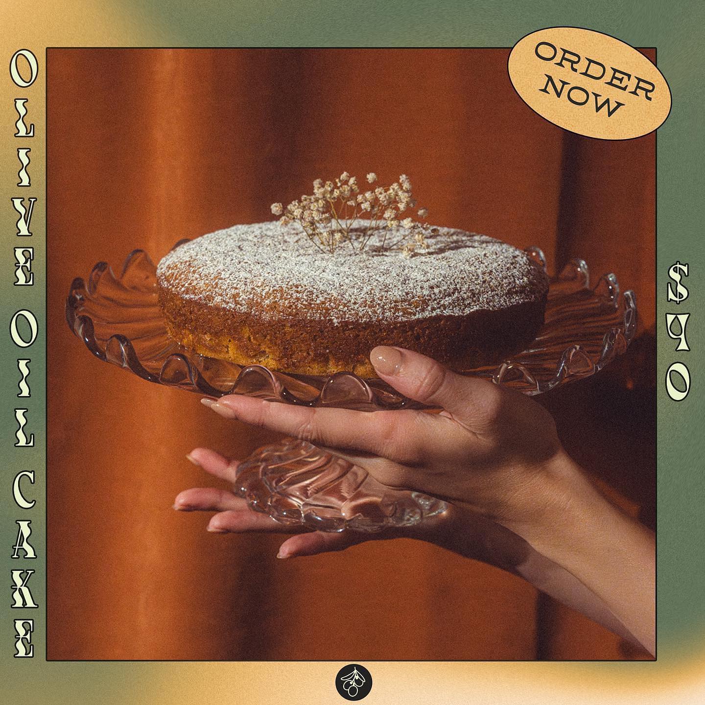 Hoonie Cakes' OG lemon olive oil cake dusted with powdered sugar, held high on a glass cake stand